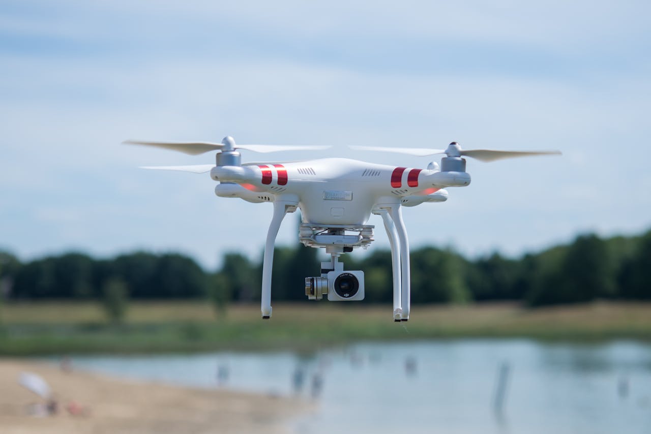 gallery-3 Aerial shot of a white drone with a camera hovering over a scenic landscape on a sunny day.