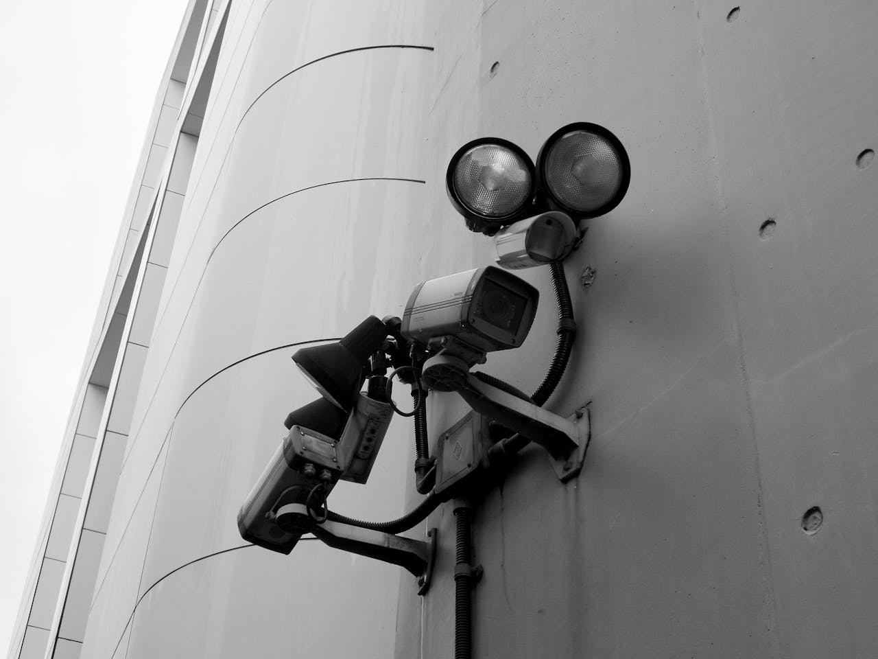 about-01 Close-up of a black and white CCTV surveillance camera with spotlights mounted on a building wall.